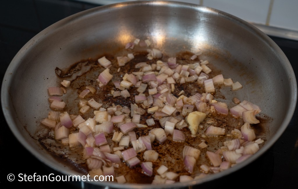 Sautéing chopped shallots in a stainless steel frying pan with a brown residue at the bottom.