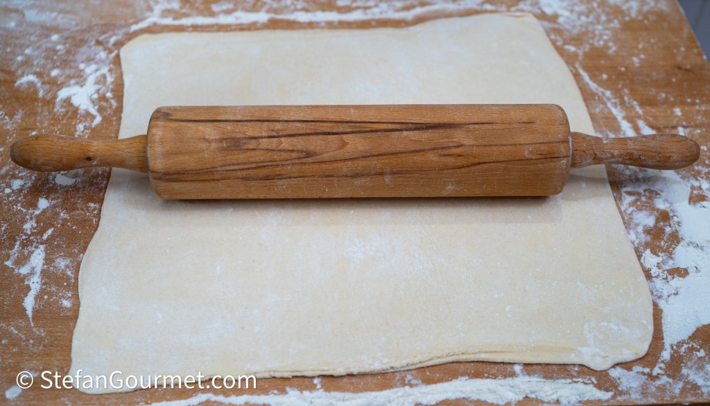 A wooden rolling pin resting on a sheet of rolled-out dough, with scattered flour on a wooden countertop.