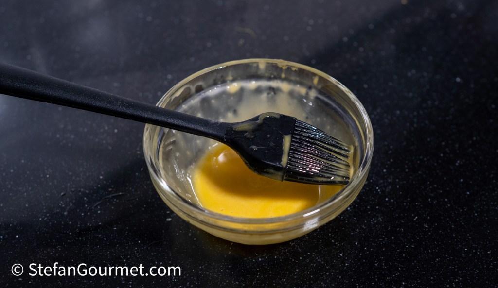 A black brush resting in a small glass bowl containing a yellow mixture on a dark surface.