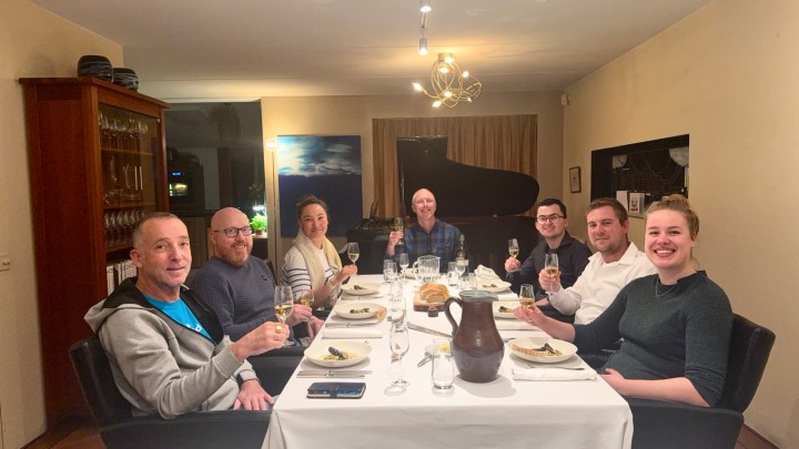 A group of six people gathered around a dinner table, enjoying a meal and toasting with drinks. The table is set with plates and glasses, and a piano is visible in the background.