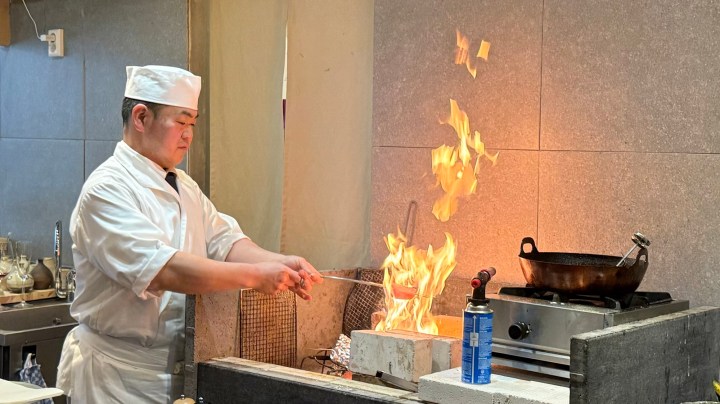 A chef in a white uniform and hat is managing an open flame in a kitchen, using a skewer to cook food over a fire.