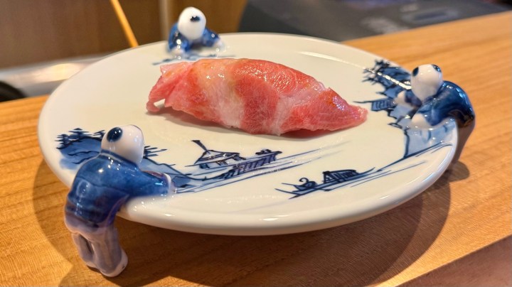 A sushi piece on a decorative plate with small ceramic figures on the edges, designed to look like they are holding the plate.