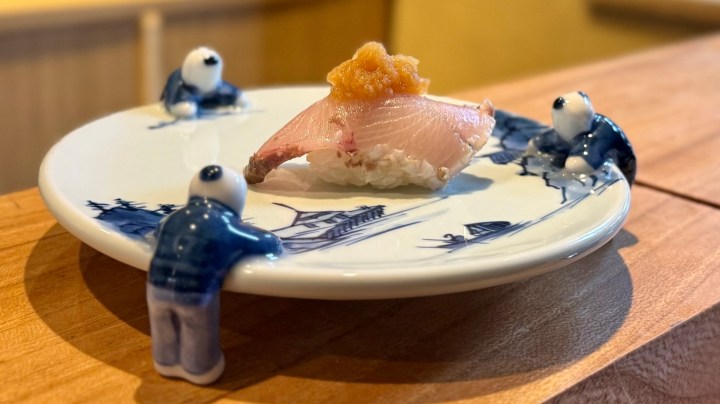 A decorative plate featuring a piece of sushi topped with a small dollop of sauce, surrounded by three playful figurines leaning against the edge of the plate, all set on a wooden surface.