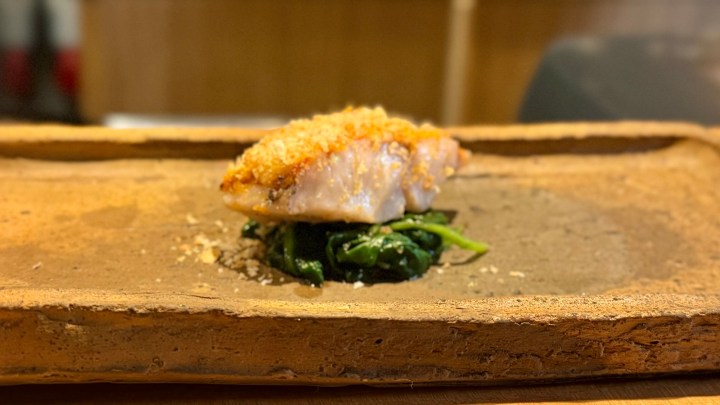 A beautifully plated piece of fish with a crispy topping, resting on a bed of sautéed greens, served on a rustic clay dish.