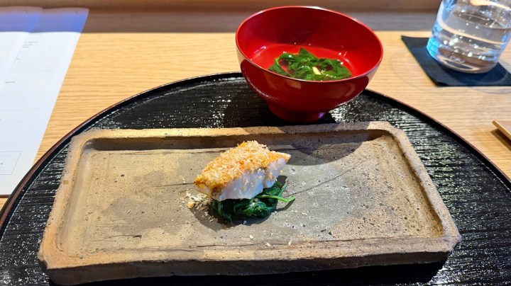 A beautifully presented dish featuring a piece of crispy fish on a bed of greens, accompanied by a small red bowl of seaweed salad.