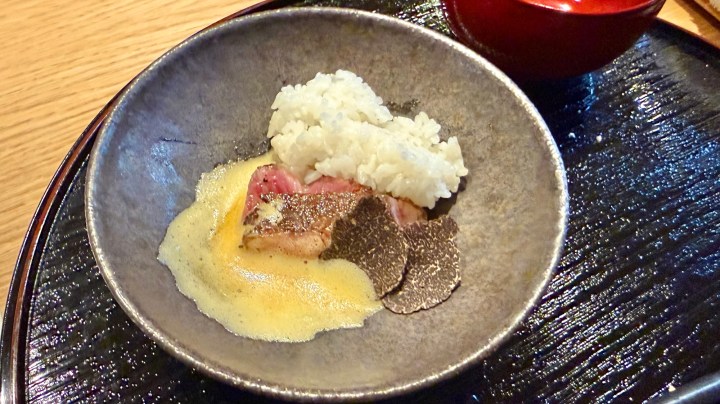 A decorative bowl containing sliced beef, a portion of rice, a creamy sauce, and shaved black truffle.