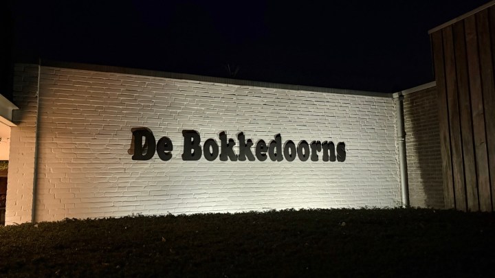 Illuminated sign reading 'De Bokkedorns' on a brick wall at night.
