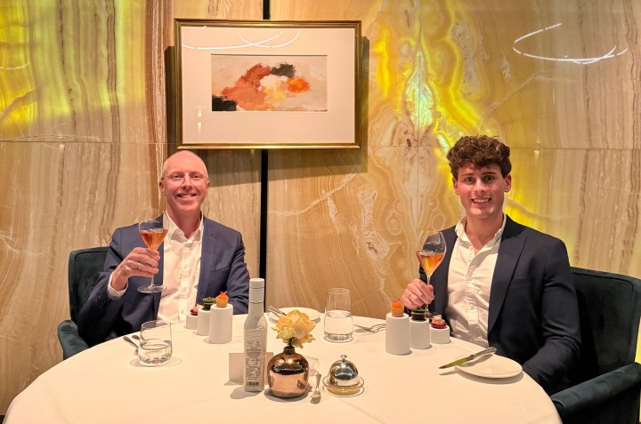 Two men sitting at a dining table, each holding a cocktail glass, smiling at the camera. The table is set with glasses, utensils, and a decorative flower, against a textured wall featuring artwork.