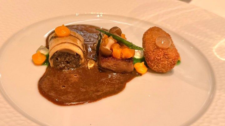 A gourmet dish featuring three elements: a rolled crepe with filling, a piece of meat topped with colorful garnishes, and a golden croquette, all presented on a white plate with a rich sauce.
