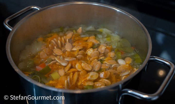 A pot of simmering soup with pumpkin seeds and vegetables, showcasing steam rising from the surface.