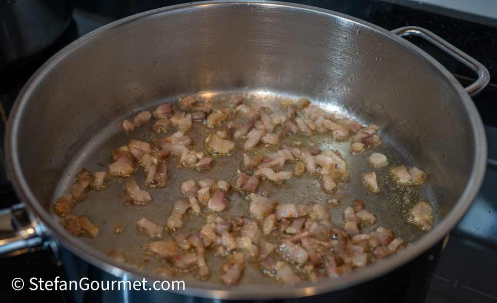 Diced bacon frying in a stainless steel pan, releasing grease and sizzle sounds.