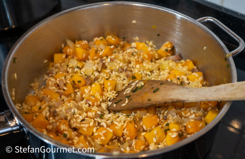 A silver pot filled with sautéed butternut squash, onions, and barley, with a wooden spoon resting inside.