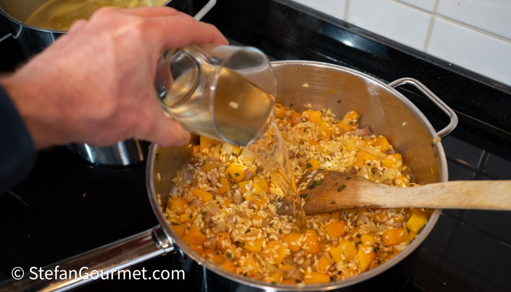 A hand pouring a glass of broth into a pot filled with rice and chopped yellow vegetables, with a wooden spoon resting inside.