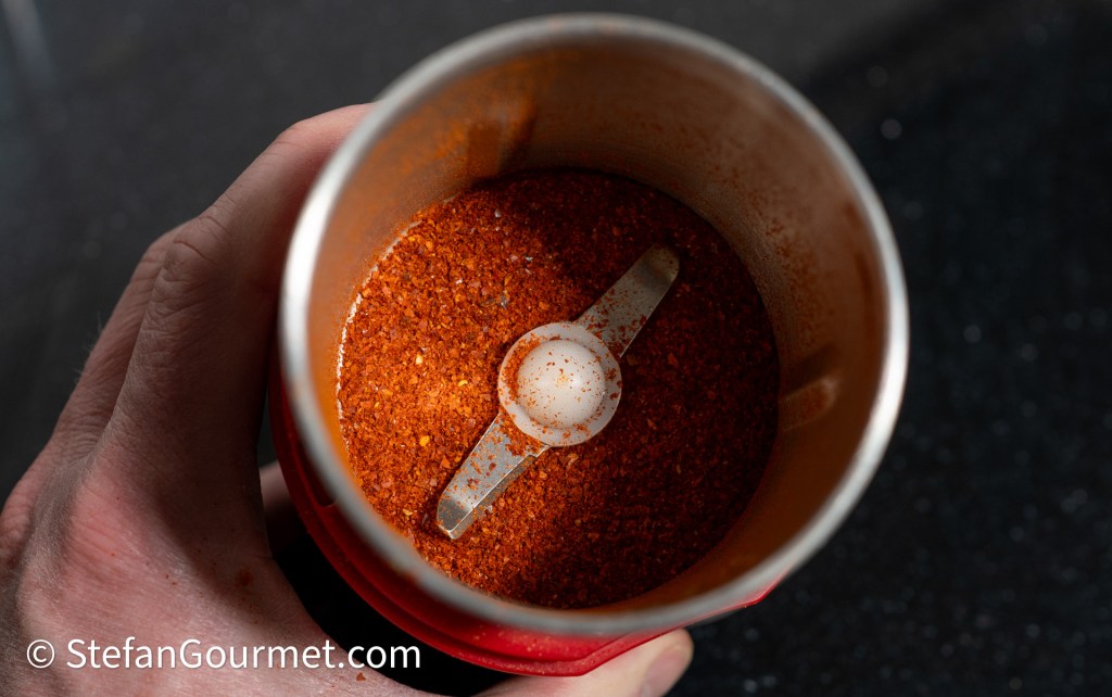 A close-up image of a spice grinder filled with orange-red spice powder and a measuring spoon inside, held in a hand.