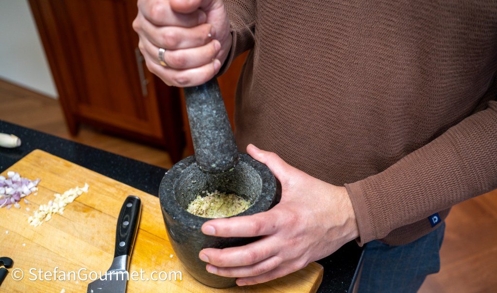 A person using a mortar and pestle to grind ingredients in a kitchen, with chopped shallots and garlic visible on a cutting board.