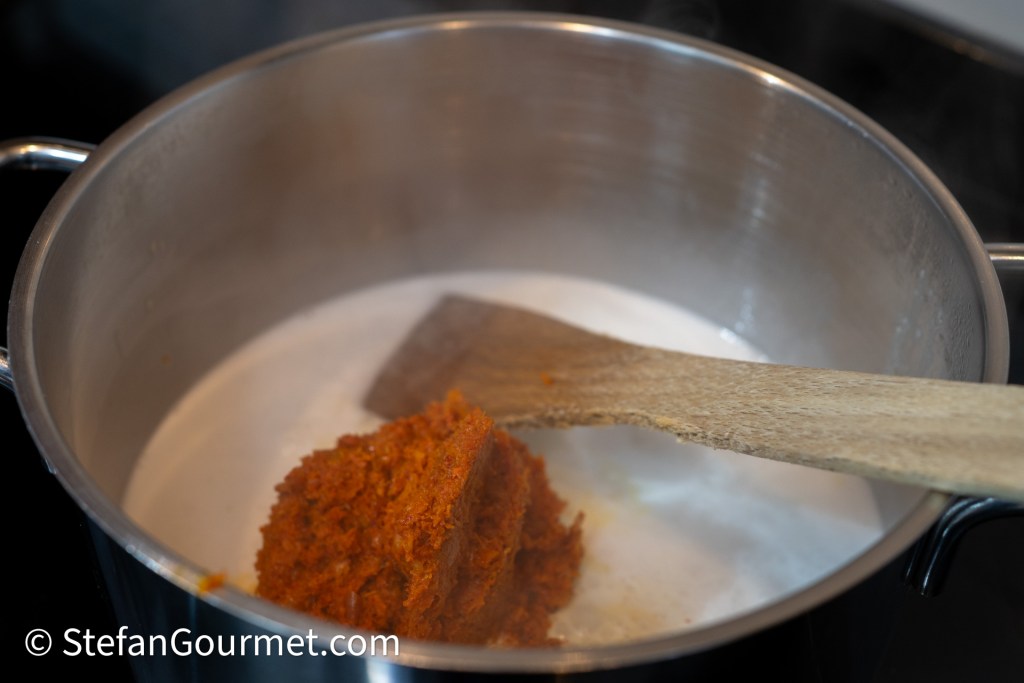 A pot on a stove with a wooden spoon, containing a dollop of orange-red cooking paste dissolving in a creamy liquid.