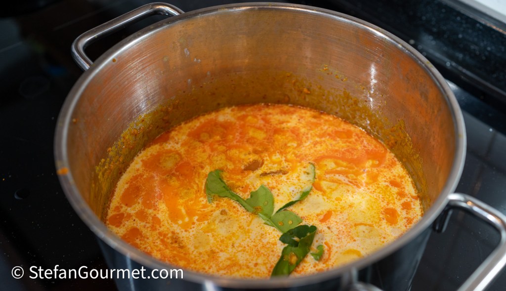 A stainless steel pot filled with a vibrant orange and creamy soup, garnished with green leaves, simmering on a stovetop.