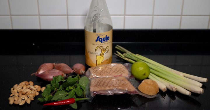 An assortment of cooking ingredients displayed on a black countertop, including cashews, shallots, fresh mint, a red chili, two pieces of grilled meat, a lime, a block of palm sugar, a bottle of squid brand fish sauce, and stalks of lemongrass.