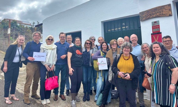 A diverse group of people posing for a photo outside a building, some holding signs that say 'Volcanic'. The scene is set in an outdoor area with cloudy skies.