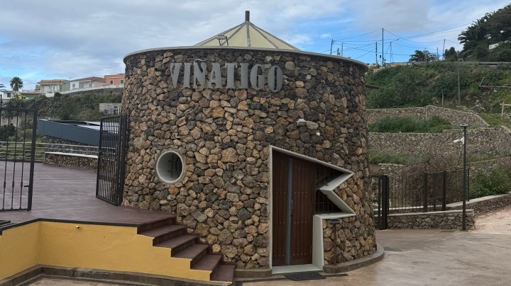 A round stone building with a conical roof, featuring the word 'VINATIGO' on the front. There are stairs leading up to an entrance door, and a fenced area is visible in the background.