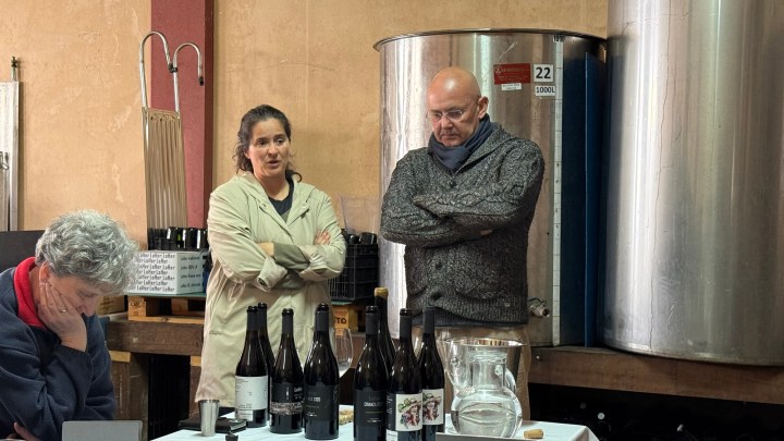 A woman in a lab coat and a man in a sweater stand near a table with wine bottles and a carafe, while another woman sits with her head down in a winery setting.