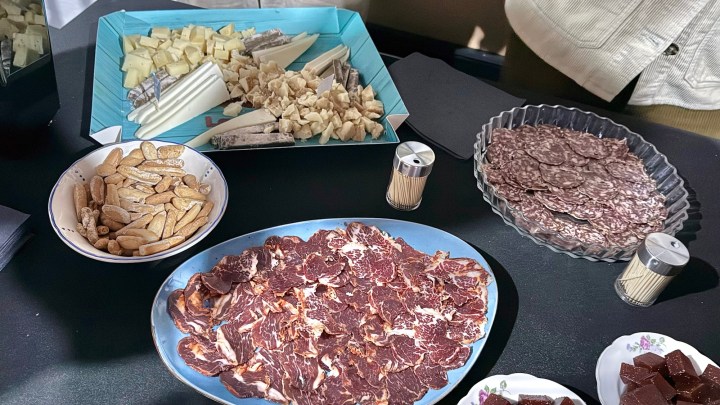 A tabletop display featuring an assortment of meats, cheeses, and snacks. There are sliced cured meats on a blue plate, various cheeses on a teal platter, and a small bowl of crackers. Additionally, there are wooden toothpicks and chocolate pieces visible.