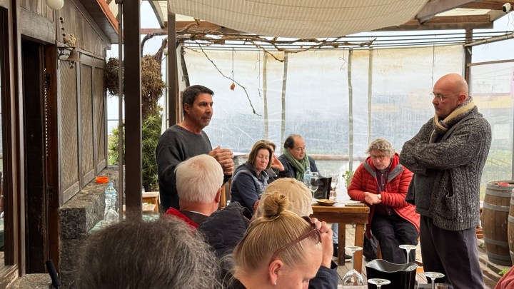 A group of people seated around tables in a covered outdoor space, with one man speaking while another listens attentively.