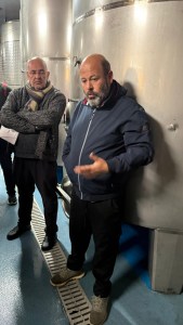 A man gesturing while speaking to a group near large metallic fermentation tanks, with another man silently listening in the background.
