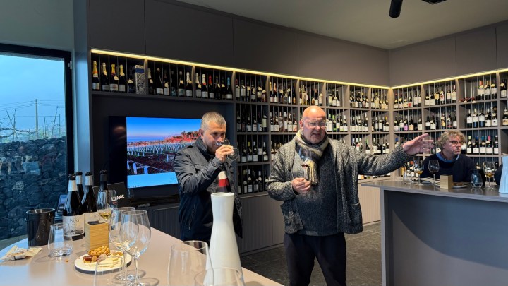 A wine tasting event featuring two men discussing wine, with shelves of wine bottles in the background and glasses on the bar.