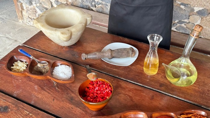 A wooden table featuring a mortar and pestle, a small plate, olive oil in a glass bottle, and an assortment of spices in wooden bowls, including salt, garlic, and chopped red peppers.