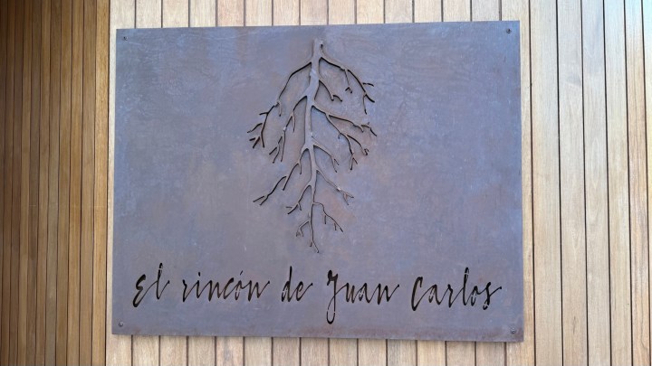 A metal sign depicting a tree root design with the text 'El rincón de Juan Carlos' engraved on it, displayed on a wooden wall.