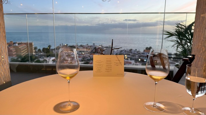 A dining table set with two wine glasses, a water glass, and a menu card, overlooking a scenic view of the ocean and city at sunset.