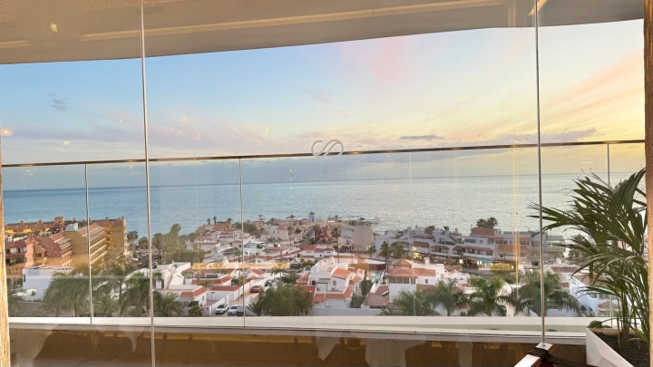 A panoramic view of a coastal town with white and colorful buildings, seen through large glass windows, overlooking the ocean at sunset.