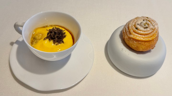 A small white cup filled with yellow dessert topped with dark garnish, alongside a round pastry with a spiral design and powdered sugar on a white plate.