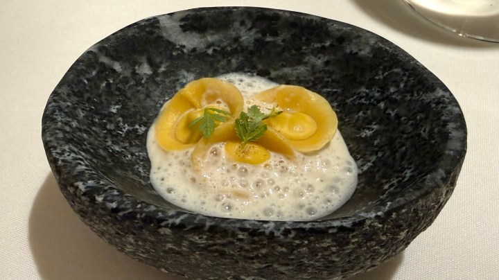 A dish presented in a dark stone bowl featuring creamy foam, yellow circular components, and a small garnish of green herb.