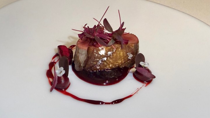 A gourmet dish featuring a round piece of meat garnished with microgreens, accentuated by a red sauce and edible flowers on a white plate.