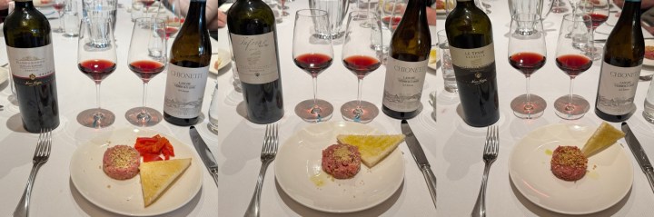 A table setting featuring four bottles of wine and several wine glasses with red wine, paired with a plate of food consisting of a ground meat dish, a slice of bread, and red peppers.
