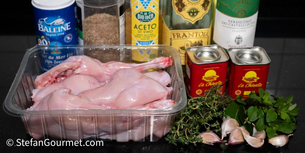 An assortment of ingredients for cooking, including rabbit pieces in a clear container, sea salt, black pepper, white wine vinegar, olive oil, and herbs like thyme and mint, along with garlic cloves and canned tomatoes.