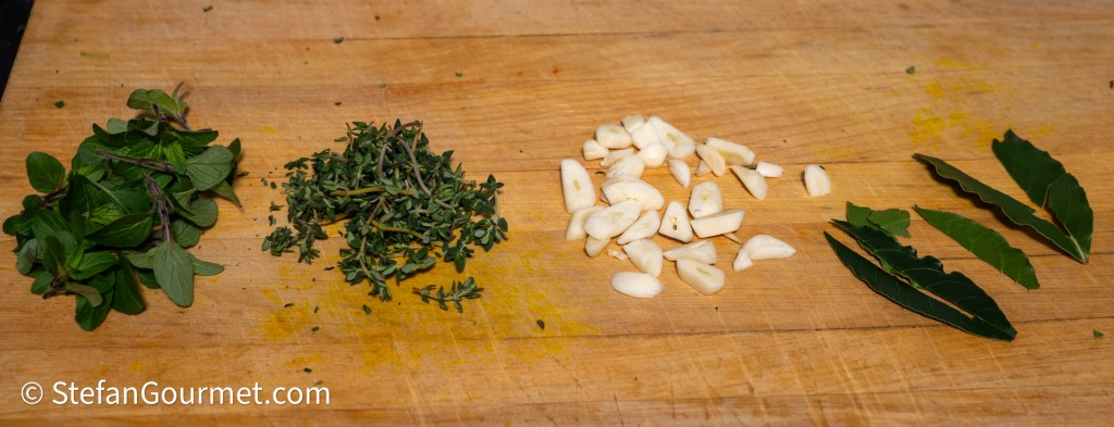 A wooden cutting board with fresh herbs: oregano on the left, thyme in the center, chopped garlic in the middle, and bay leaves on the right.