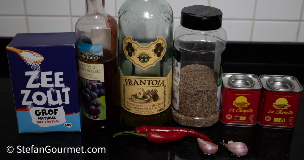 A collection of cooking ingredients on a black countertop, including a box of sea salt, a bottle of red wine vinegar, an olive oil bottle, a jar of ground cumin, a red chili pepper, two cloves of garlic, and two cans of paprika.