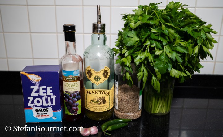 A selection of cooking ingredients including a box of sea salt, bottles of red wine vinegar and olive oil, a jar of ground black pepper, fresh parsley in a glass, two jalapeño peppers, and a couple of garlic cloves on a dark countertop.