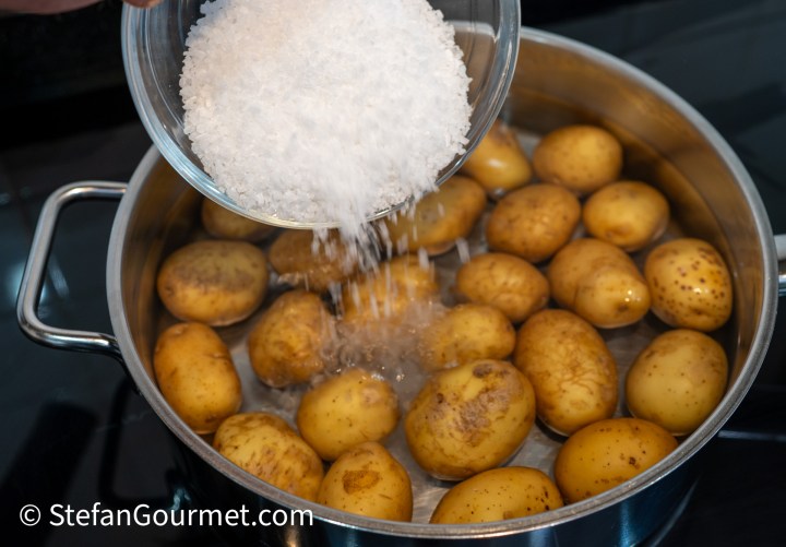 A person pouring coarse salt from a bowl into a pot of boiling potatoes on a stovetop.