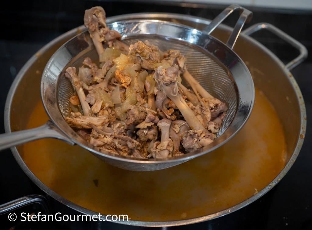 A colander holding cooked chicken bones and vegetables over a pot of broth.