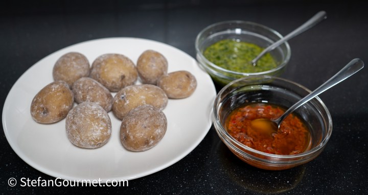 A plate of boiled potatoes with a dusting of flour, accompanied by two small bowls of sauces: one green and one red.
