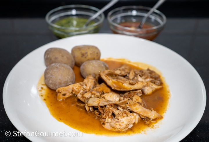 A plate of chicken in a flavorful sauce, served with small boiled potatoes and two bowls of dipping sauces, one green and one reddish-brown.