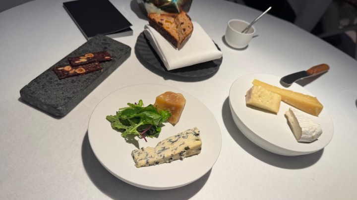 A table set with various cheeses, including blue cheese, creamy cheese, and hard cheese, accompanied by greens and a honey-like gel. In the background, there is a plate of baked goods and a cup.